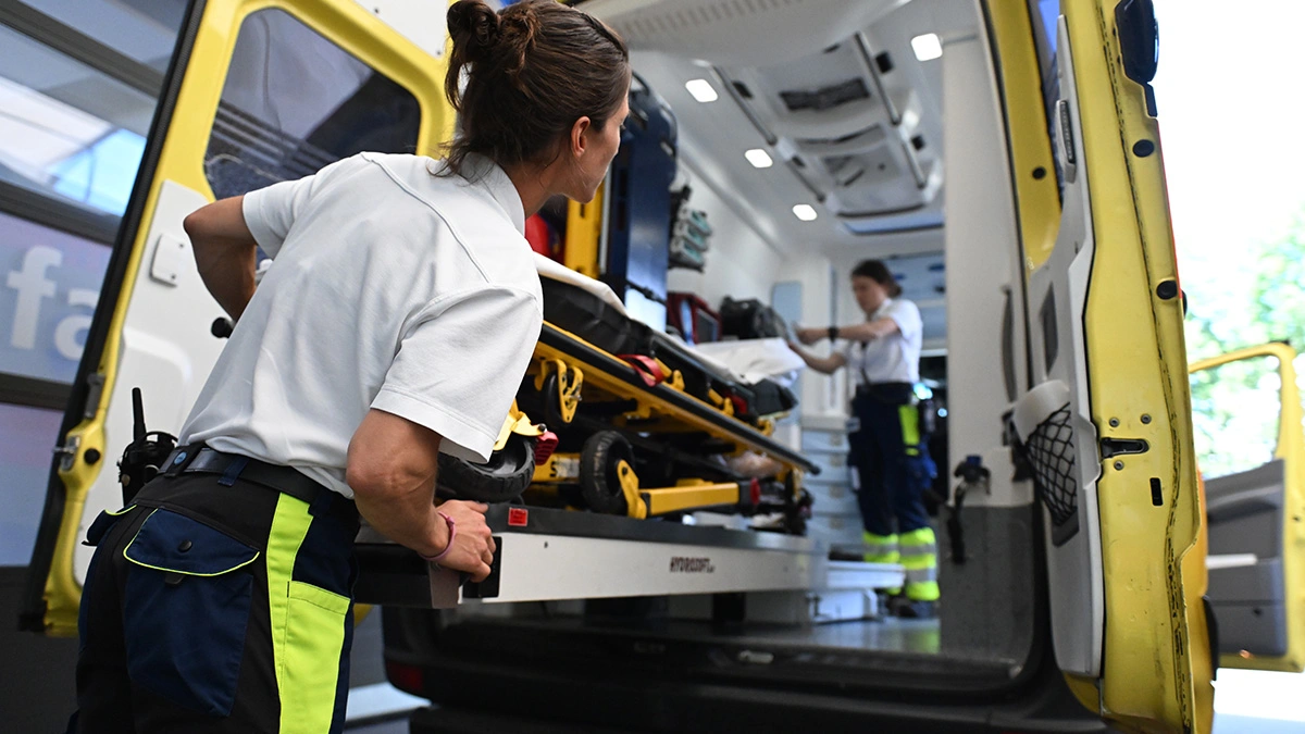 Rettungssanitäterin lädt Trage in Krankenwagen, während Kollege im Inneren des Rettungswagens Ausrüstung vorbereitet.
