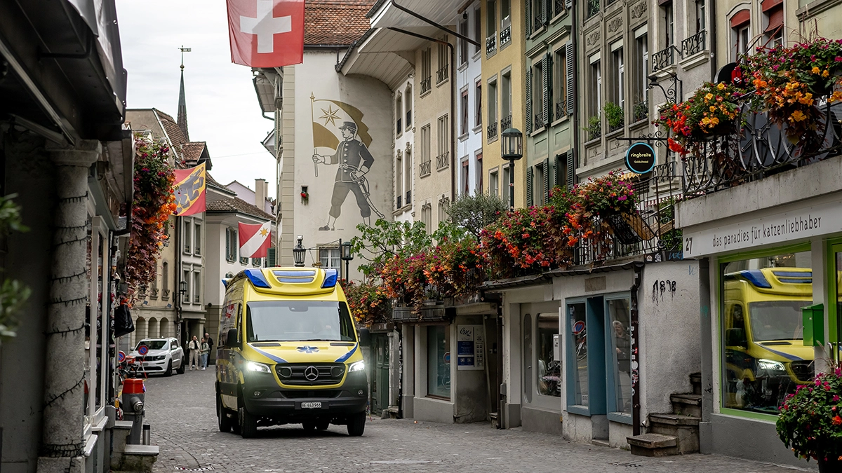 Rettungswagen fährt durch historische Altstadt mit Schweizer Flagge und blumengeschmückten Fassaden, Rettungsdienst im urbanen Einsatz.
